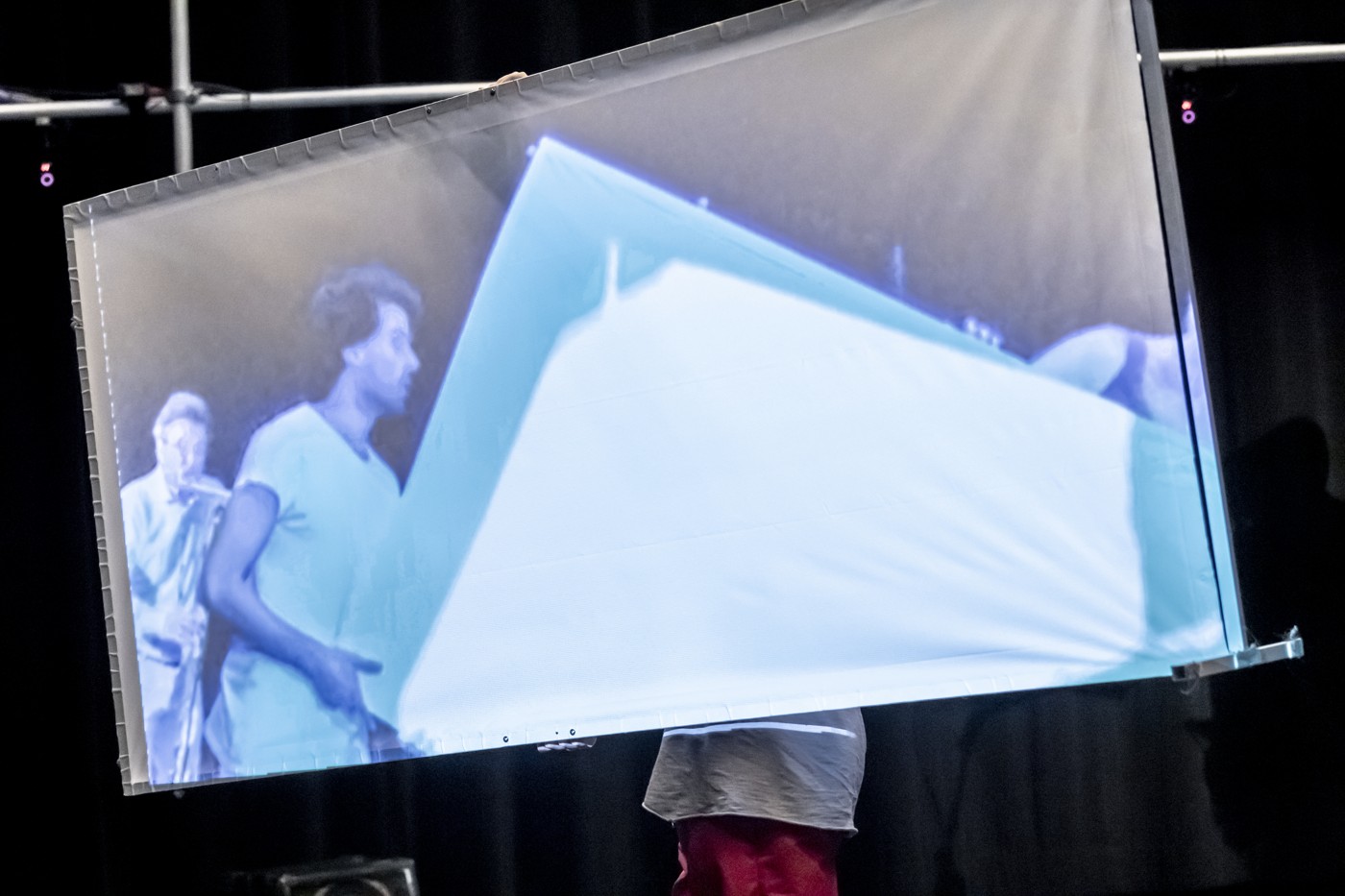 A performer carrying a tilted screen displaying a remote space.