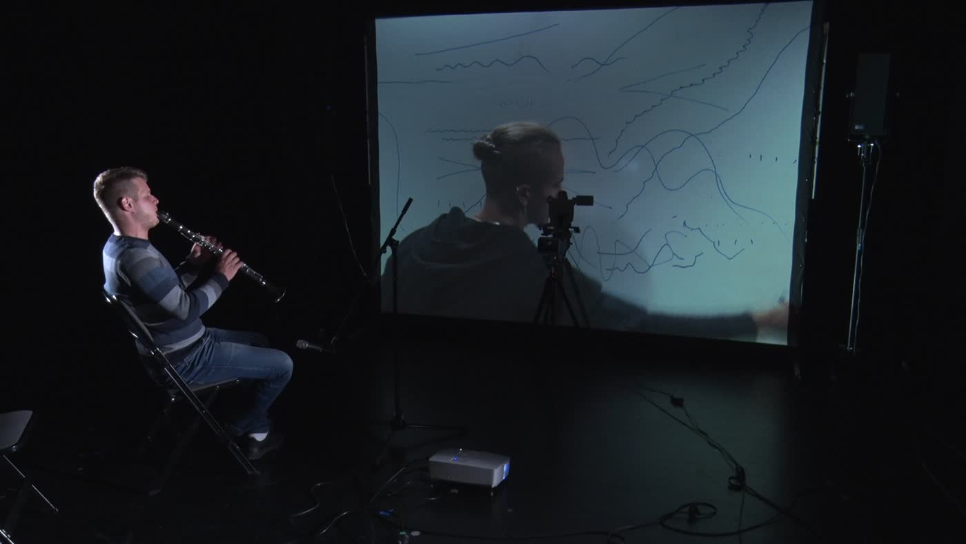 Clarinet player seated beside a camera and a projection of another participant.